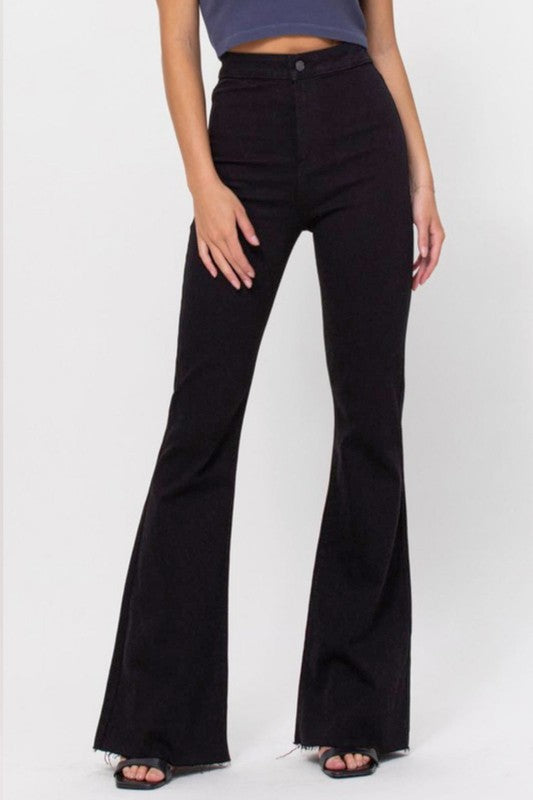Woman wearing high-waisted, black flared jeans and a dark crop top, standing against a plain white background with her hands resting at her sides. Only her body is visible from shoulders down.