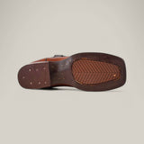 The image features the sole of a Grisly Tan - Square Toe boot, made of premium leather with stitching and small perforations. This Western-style shoe has a textured grip and rectangular heel, set against a plain light gray background.
