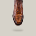 A brown shoe with crocodile skin texture is displayed against a plain background. The Exotic American Alligator Tail - Fawn boot, featuring a handcrafted design, faces the viewer toe-first to emphasize its detailed pattern and stitching.