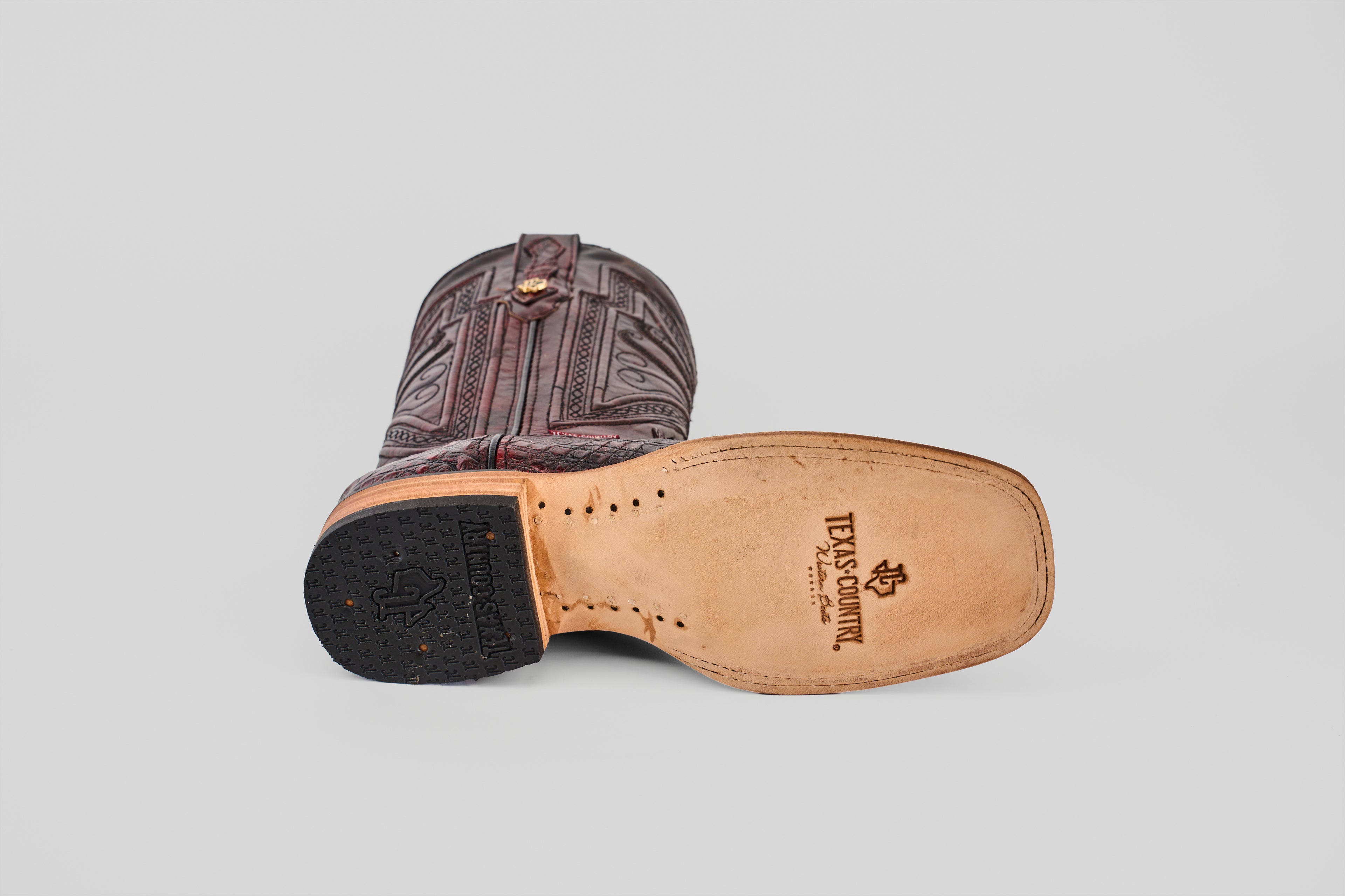 A single Exotic Caiman Belly - Black Cherry - Square Toe boot lies on its side, revealing intricate leather patterns, a light sole stamped with the Texas Country logo, and a heel with a black rubber star design.