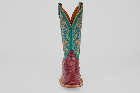 The Rusty - Wine - Square Toe cowboy boot features a wine-hued ostrich leather foot and a tall teal shaft with detailed yellow and white stitching, displayed facing forward on a plain light background.
