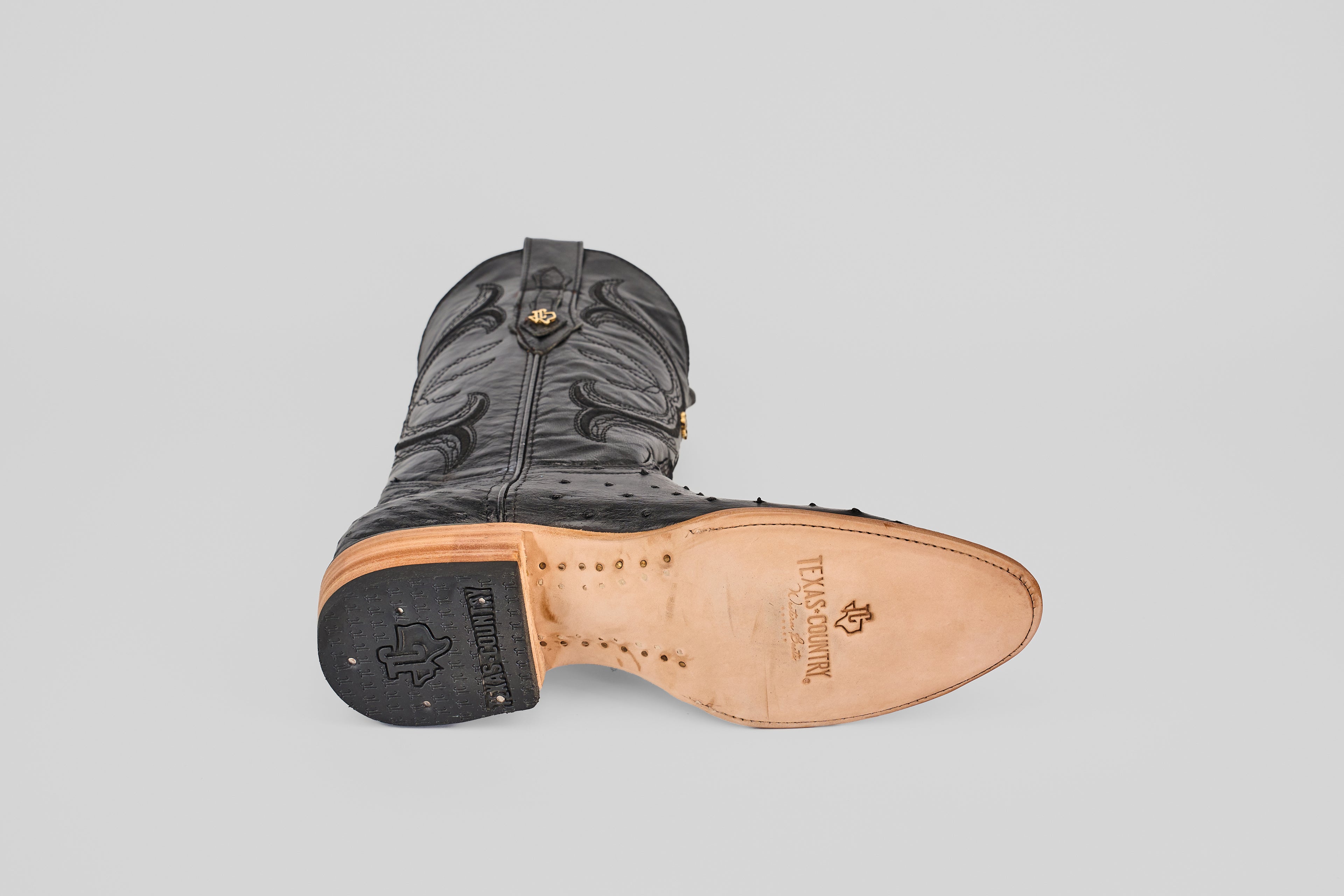 The Austin - Black - Roper Toe boot rests on its side, highlighting intricate stitching, a tan sole stamped “TEXAS COUNTRY,” and a logo on the black heel. The scene is set against a light gray background.