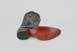 A single Tanner - Black - Round Toe cowboy boot rests on its side, highlighting detailed stitching on the upper and a smooth brown Python Negro Oval leather sole with the Black Star by Justin logo.