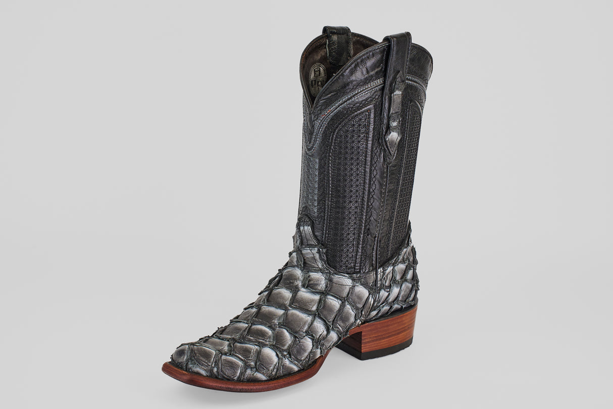 The Conor - Newspaper boot features a black, narrow square toe with a scale-like lower pattern reminiscent of fish leather, a smooth leather upper, pull straps, and a brown wooden heel, all showcased on a white background.