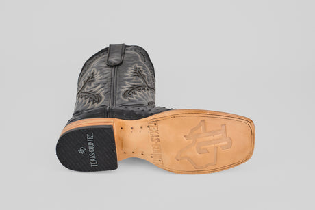 The Ostrich Black - Embossed Leather - Square Toe (E424) boot is shown on its side, highlighting decorative stitching, a tan sole with TEXAS COUNTRY and logo engraving, and a black heel printed with TEXAS COUNTRY in white.