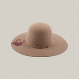 The 4x - Natural - TC Hats is a wide-brimmed, light brown felt Western hat with an open crown, a small decorative band, and a maroon tag on the brim, shown against a plain, light background.