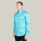A man in a Men’s Single Pocket Logo Modern Fit Stretch Dress Shirt - Turquoise (CTL9829) and black pants stands against a plain beige background, slightly facing left with a neutral expression.