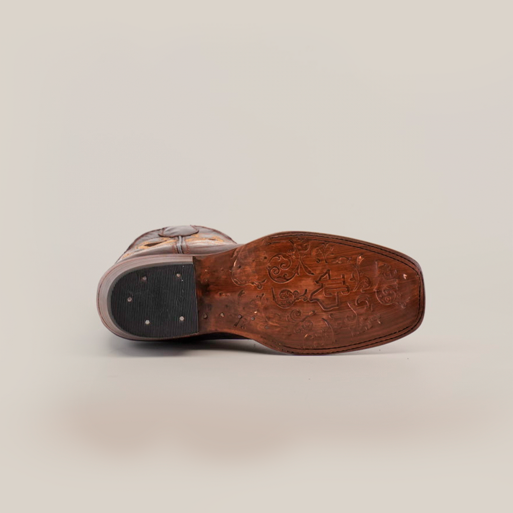 The image shows the underside of a Western Stitch Tabaco-Short Shaft-Narrow Square Toe boot. Superior leather quality is clear in the intricate sole engravings, and a black rubber heel with metal studs stands out against a plain white background.